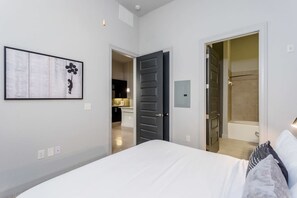 A bedroom with a bed, a door leading to a bathroom, and a kitchen visible through an open door. - Peony At Millennium - Elegant 2 BDR in Houston (Houston)