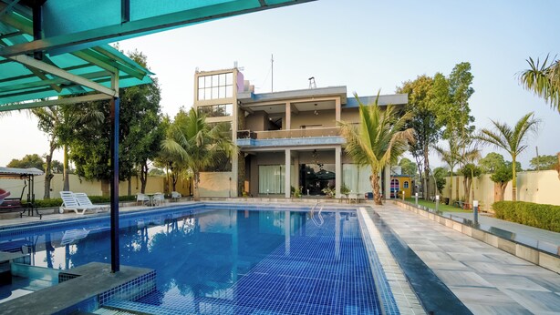 A swimming pool with a tiled deck, surrounded by palm trees and a modern building with balconies. - Polki Farm Resort Jawai -Sumerpur (Sumerpur)
