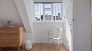A room with a sloped ceiling, a wooden dresser, a white chair, a window with white frames, and a door. - Serene Cottage Nr the Waterfront, Topsham (Topsham)