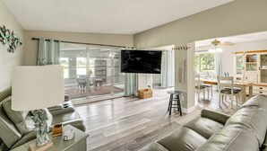 A spacious living room with a flat-screen TV, leather sofas, a dining area with chairs, and a large sliding glass door leading to an outdoor patio. - Saltwater Pool | Coastal Family Retreat (Port Charlotte)