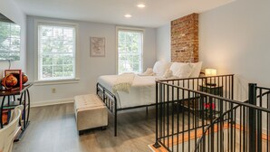 Ein Schlafzimmer mit einem Bett, zwei Fenstern, einer Ziegelfassade, einem Nachttisch mit Lampe und einem kleinen Hocker.