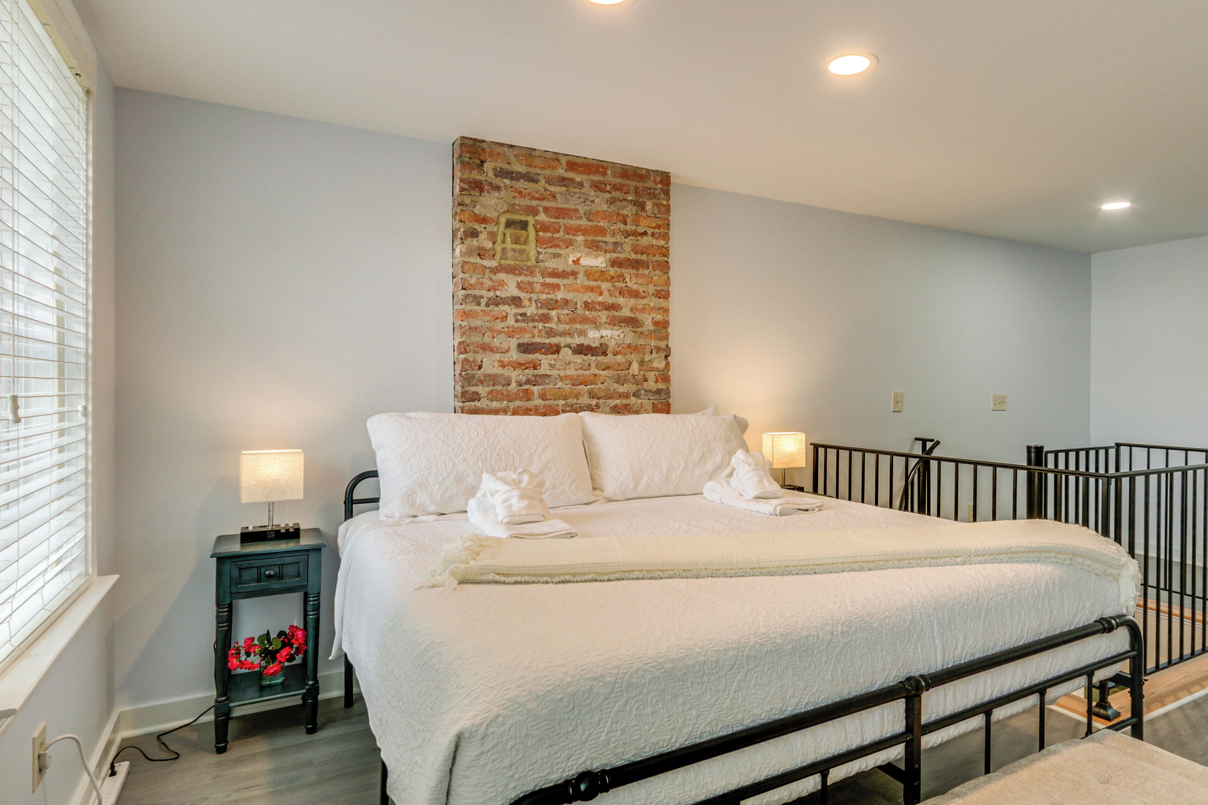 A bedroom with a brick accent wall, a bed with white linens, two bedside lamps, and a nightstand with a red flower arrangement.