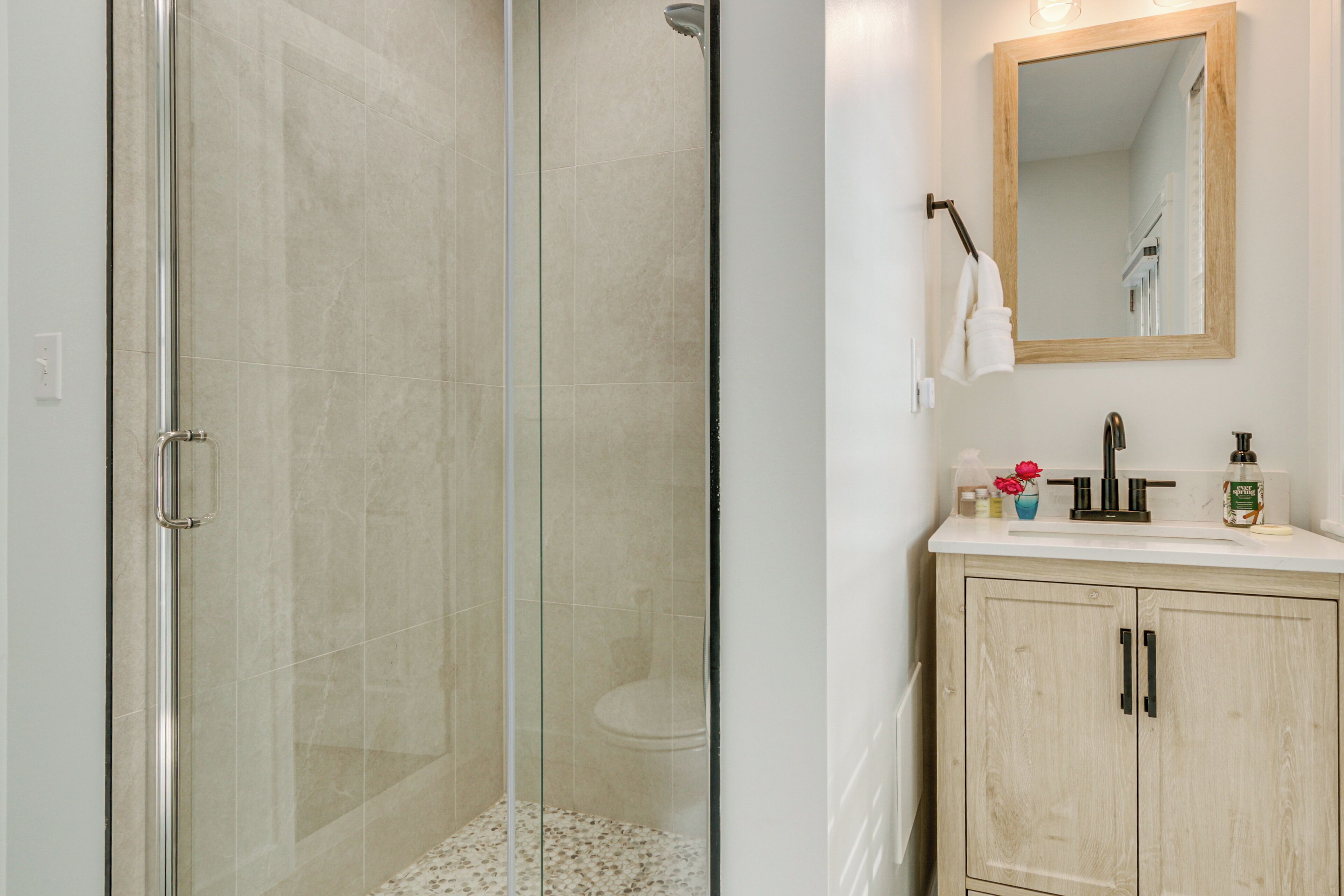 Un baño moderno con ducha envuelta en vidrio, un lavamanos blanco con espejo arriba y un mueble de madera abajo.