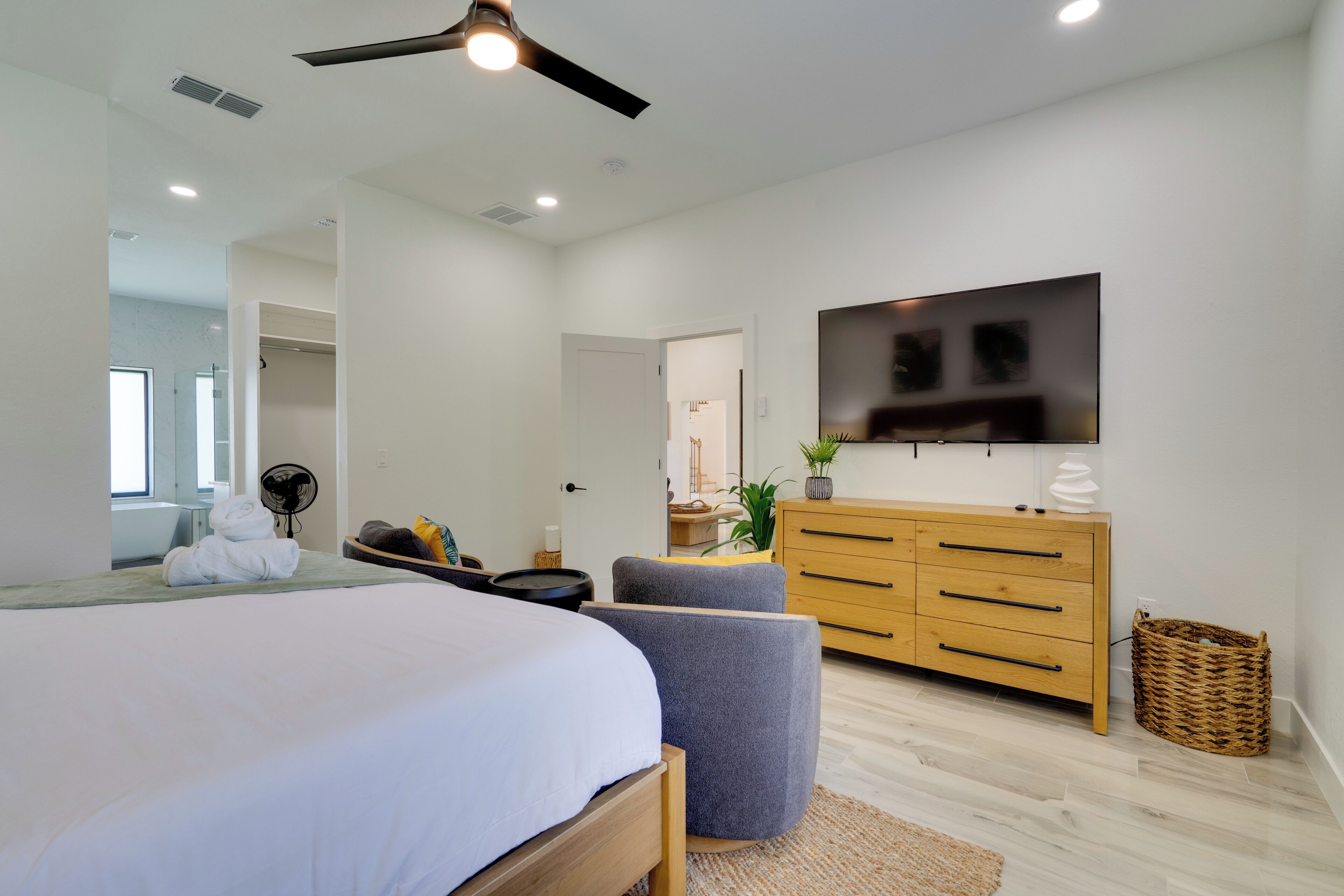 Ein modernes Schlafzimmer mit einem großen Bett, einem Deckenventilator, einem an der Wand montierten Fernseher und einer Holzkommode.