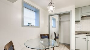 Een eetruimte met een glazen tafel en stoelen, een kroonluchter, een raam met zonwering, en een keuken met kasten.
