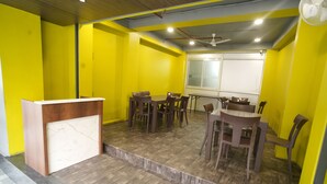 A room with yellow walls, a wooden reception desk, multiple tables and chairs, and a ceiling fan. - The Executive Inn (Siliguri)