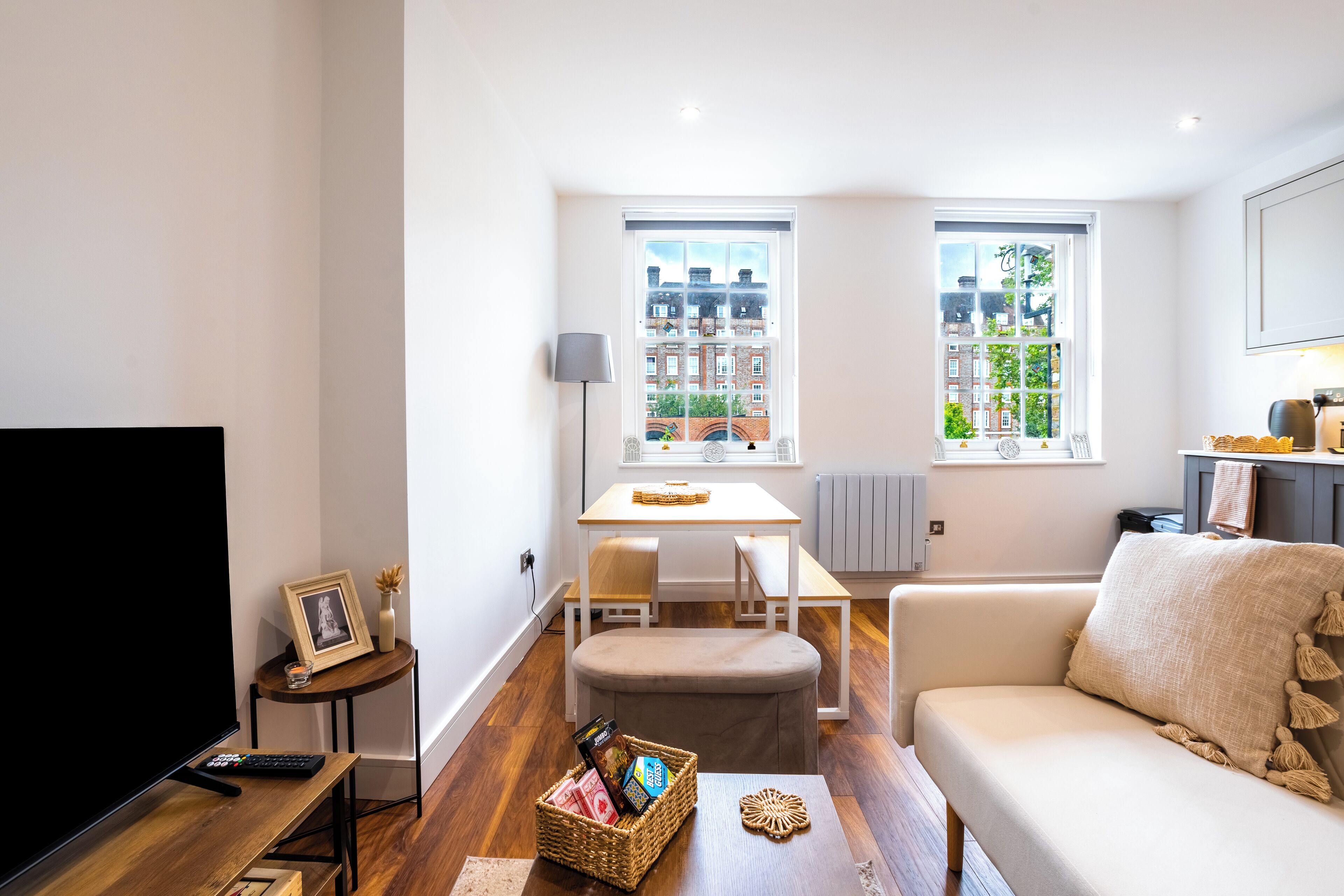 Uma sala de estar moderna com sofá, TV, área de jantar com mesa e cadeiras, e amplas janelas com vista para edifícios.