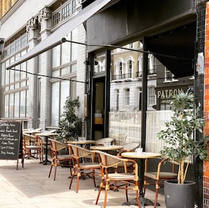 An outdoor cafe with wicker chairs and tables, a chalkboard sign advertising food and drink, and a view of a building with a sign reading "PATRON".