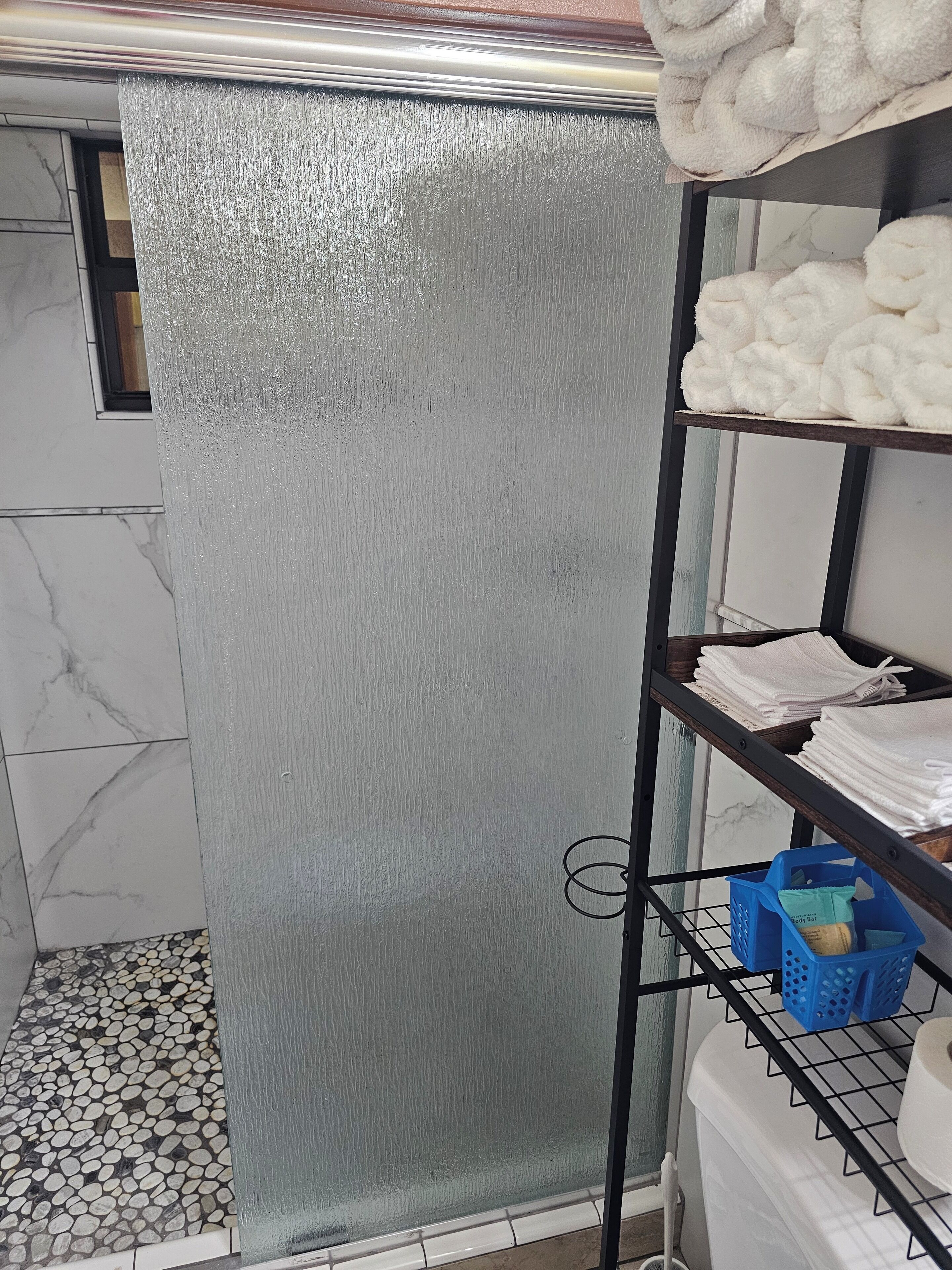 A bathroom with a shower area, a tiled floor, and a shelf with towels and toiletries.
