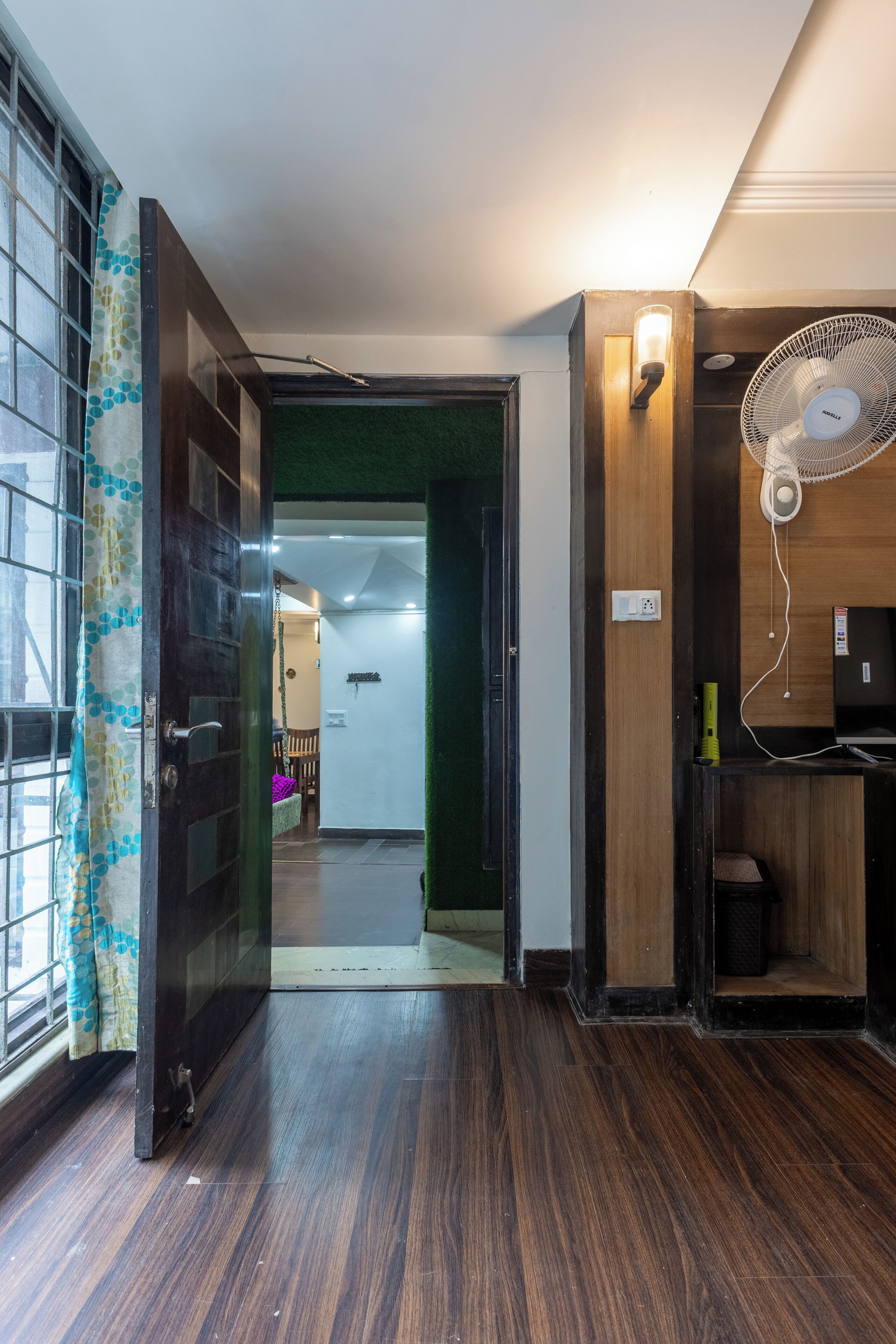 Um corredor interno com piso de madeira, porta de vidro com cortina, ventilador fixado na parede e porta verde que dá para outro quarto.