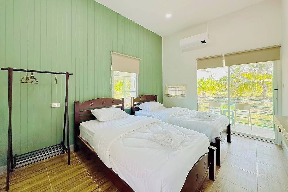 A hotel room with two beds, a wooden floor, a window with a view of greenery, and an air conditioning unit.