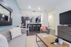 A modern living room with a sofa, coffee table, and a TV. A kitchen with a bar area is visible in the background.