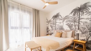 A bedroom with a bed, a ceiling fan, a bedside table, a nightstand, a woven bench, and a wall with a tropical mural. - Stylish Urban Escape – 2-Bedroom Apartment with Rooftop Pool (Estepona)