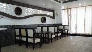 A restaurant with a modern interior, featuring a curved wall with decorative cutouts, a row of tables and chairs, and a large window with curtains.