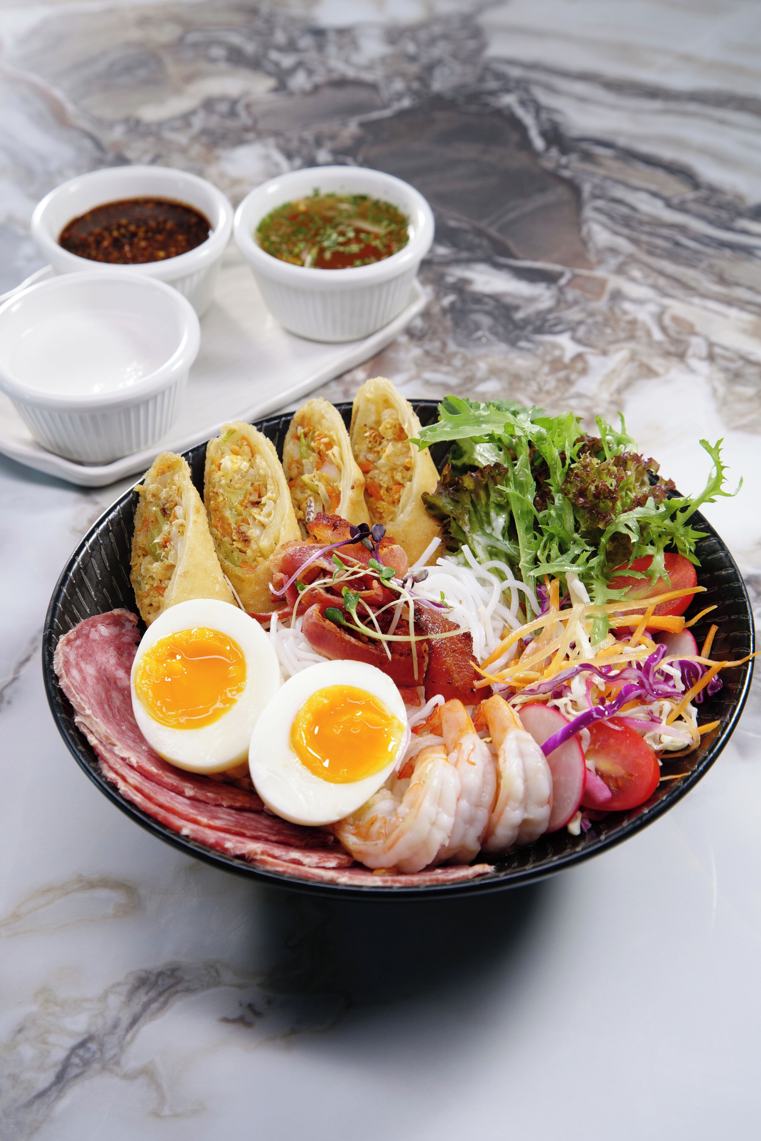 A bowl of food with various ingredients and two small bowls of sauce.