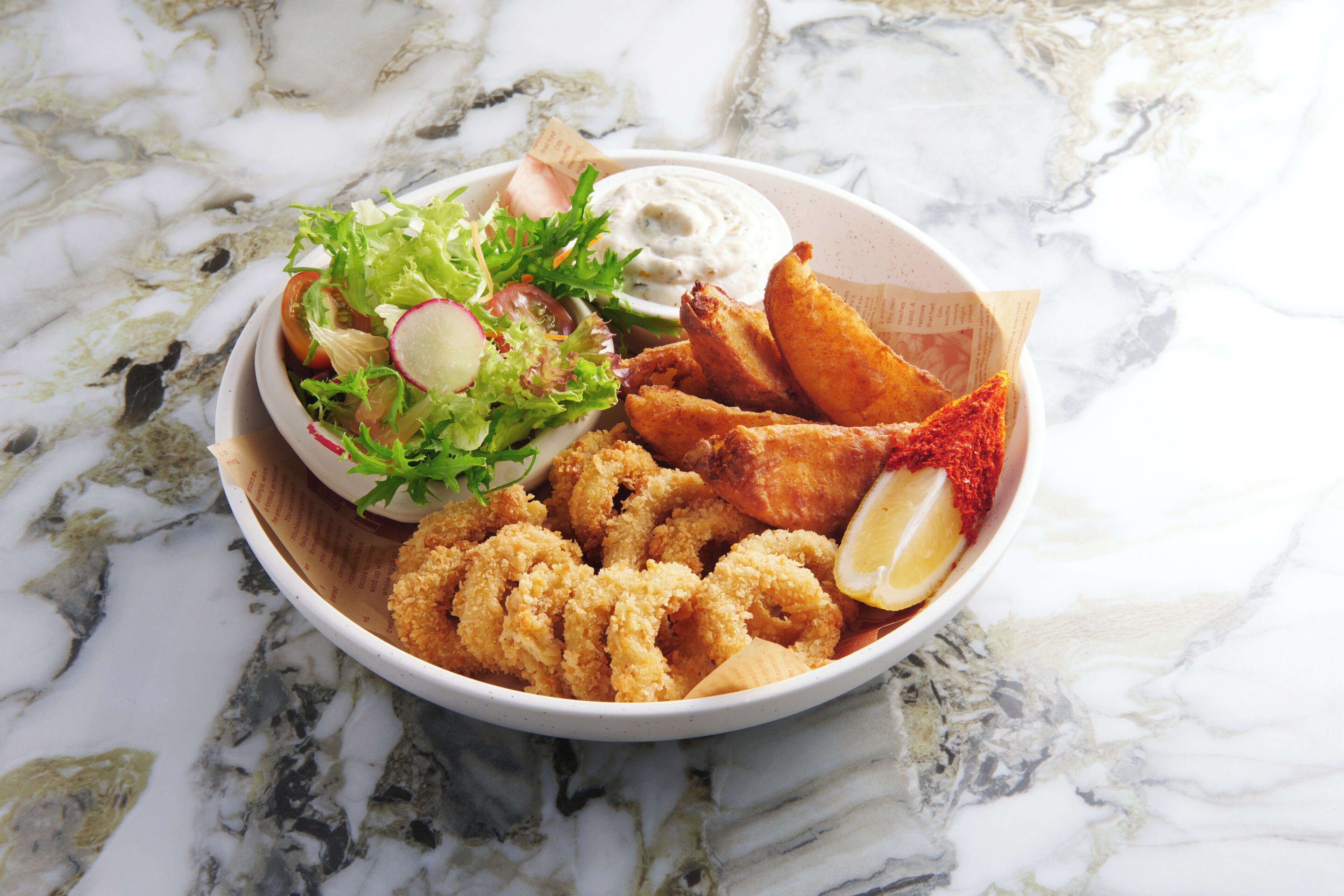 A bowl of seafood including fried calamari, lemon wedges, and a side salad with radish and lettuce on a marble surface.