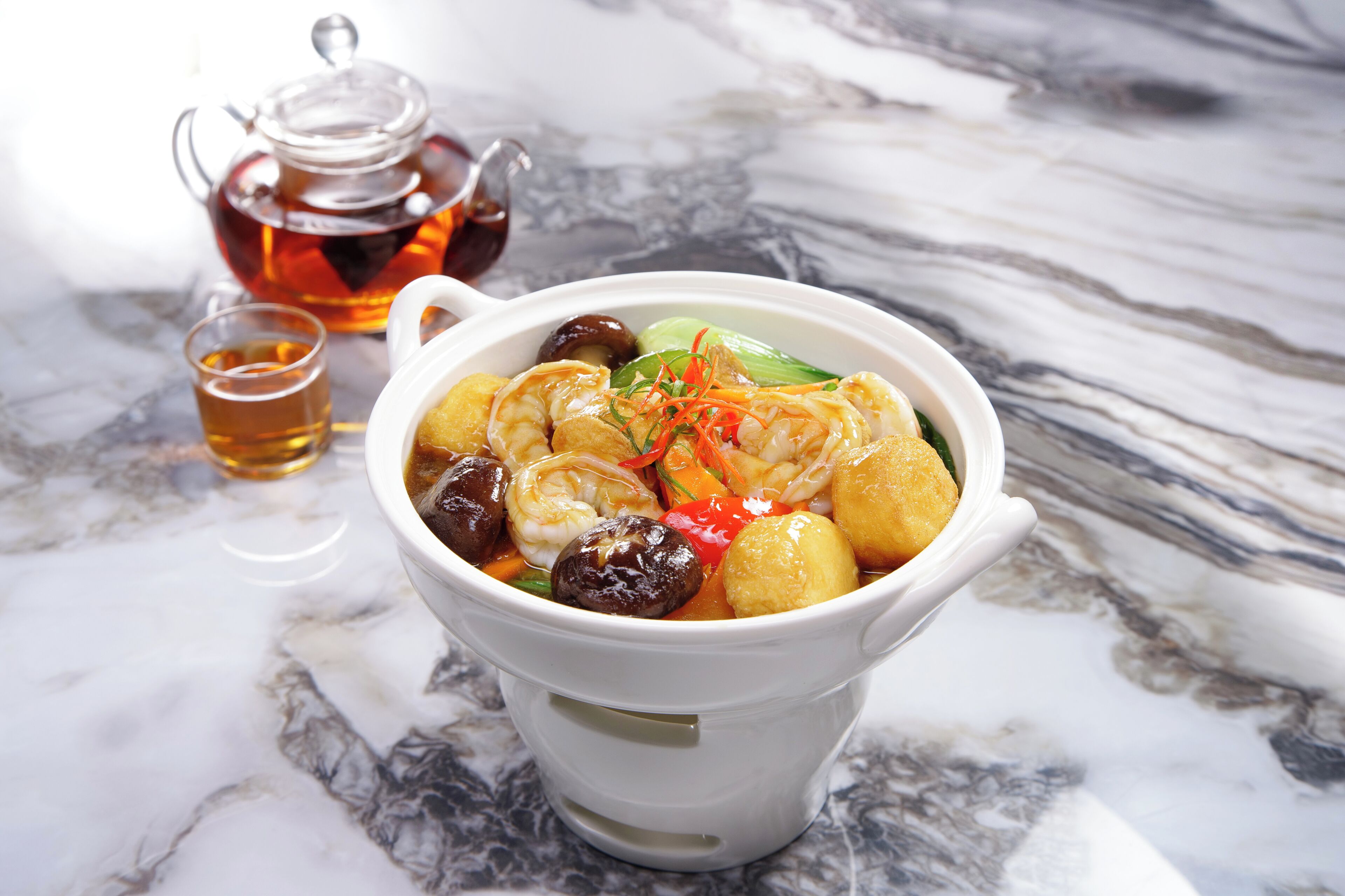 A bowl of soup with various ingredients, a teapot, and a cup of tea on a marble surface.