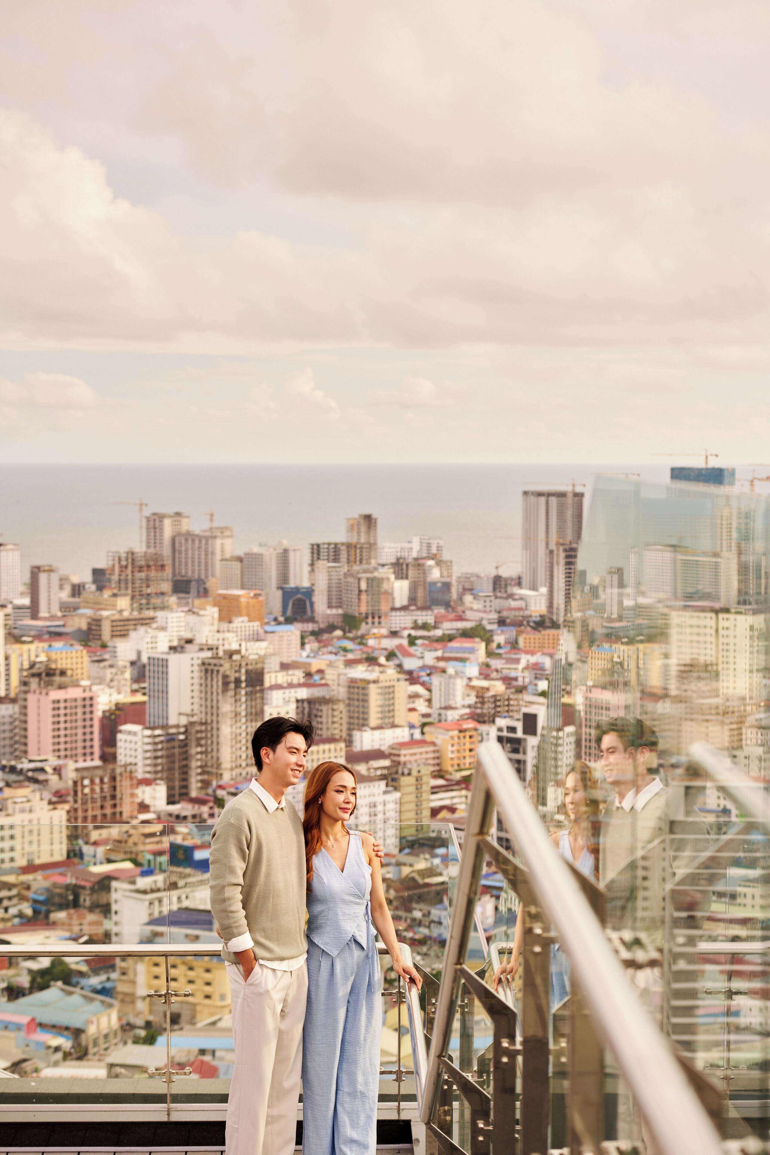 Un couple se tient sur un balcon, d’où l’on peut admirer un paysage urbain avec de hauts immeubles et une vue sur la mer.