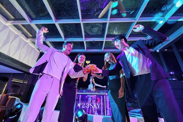 A group of people toasting with drinks in a modern, well-lit room with a glass ceiling and a visible bar area.