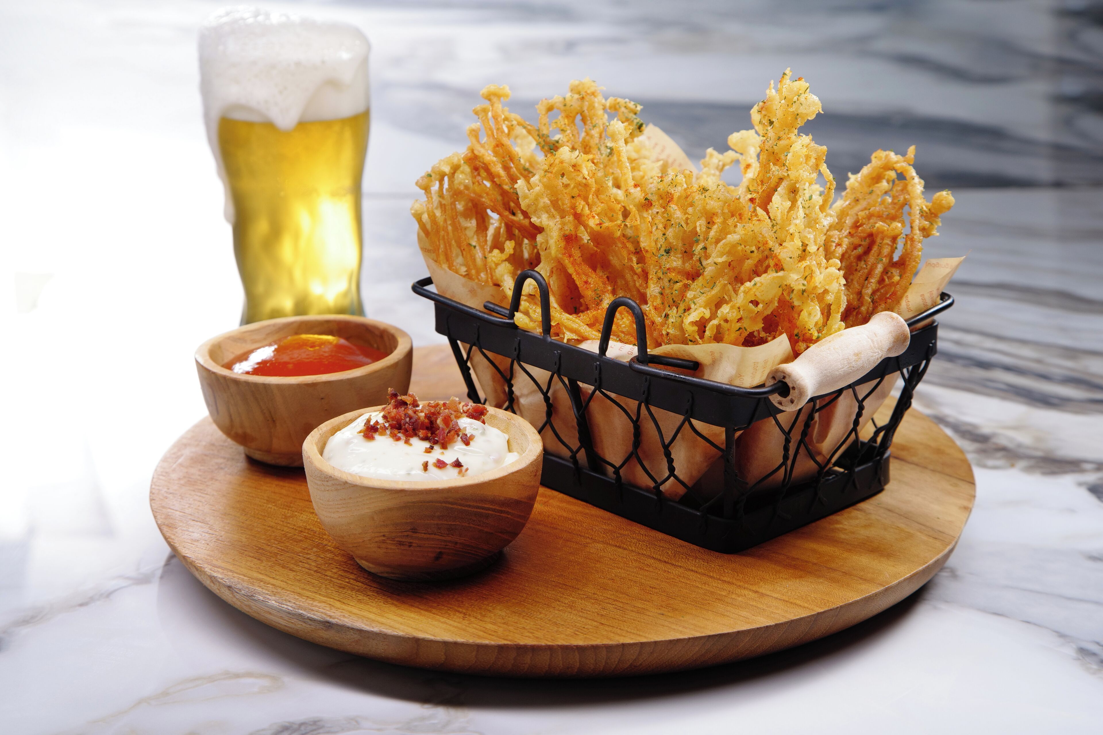 A wooden tray with a basket of fried food, a bowl of sauce, and a glass of beer on a marble surface.