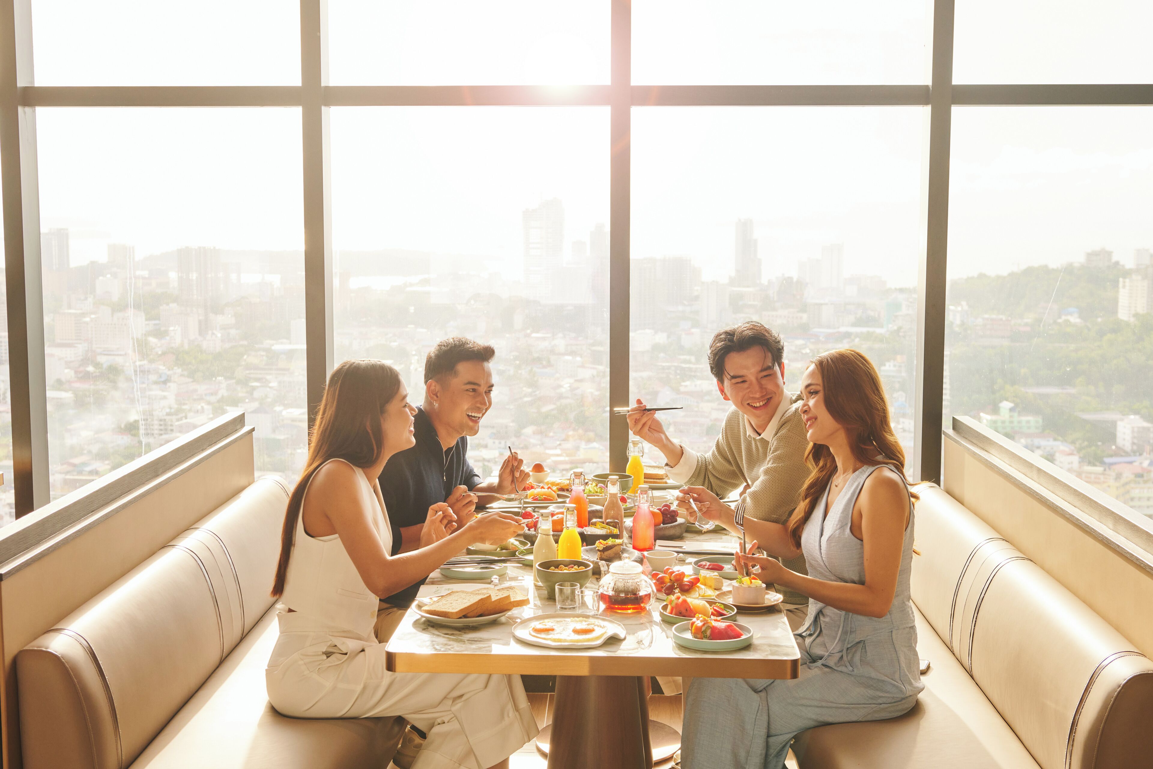 Un groupe de personnes est réuni autour d’une table pour dîner, avec une vue sur la ville. Elles se trouvent dans une salle dotée de grandes fenêtres et d’un décor moderne.