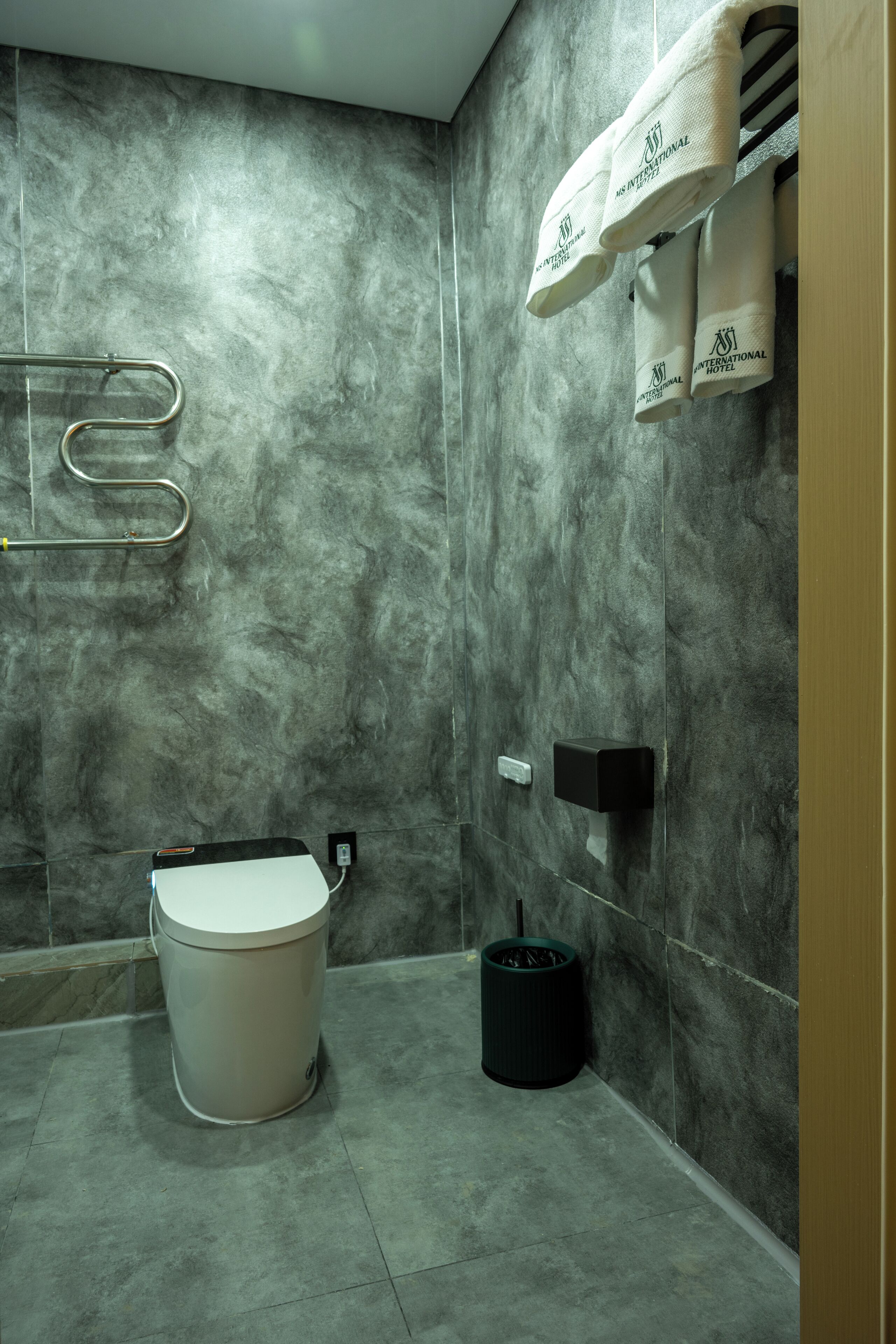 A bathroom with a marble wall, a toilet, a trash bin, and towels hanging on a rack.