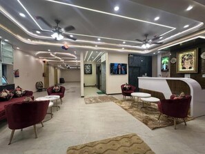 A modern hotel lobby with a reception desk, seating area with red chairs and white tables, and a ceiling with recessed lighting and a fan. - Hotel Vrinda Shridhar (Vrindavan)