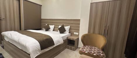 A modern hotel room with a large bed, a wooden wardrobe, a small bedside table, and a patterned armchair.