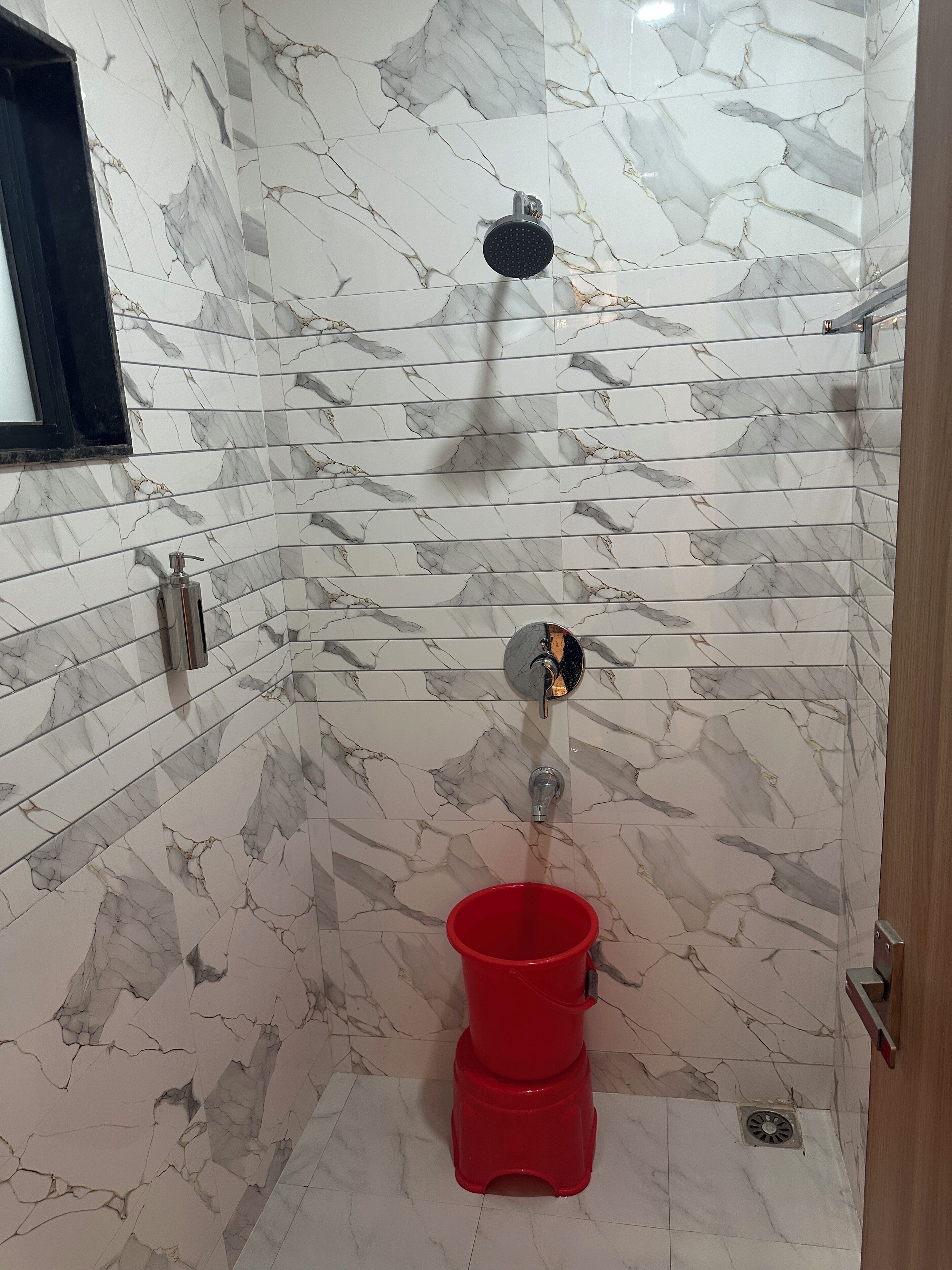 A bathroom with a shower, a red bucket, and a marble-patterned wall.