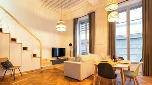 Un salon moderne avec un canapé, une table à manger et des chaises. On y trouve également un téléviseur, un escalier et de grandes fenêtres avec des rideaux.