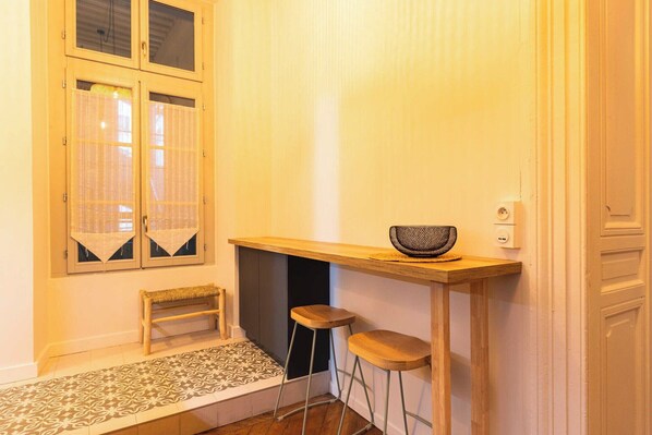A small kitchen area with a wooden bench, two stools, a small table with a bowl, and a window with curtains.