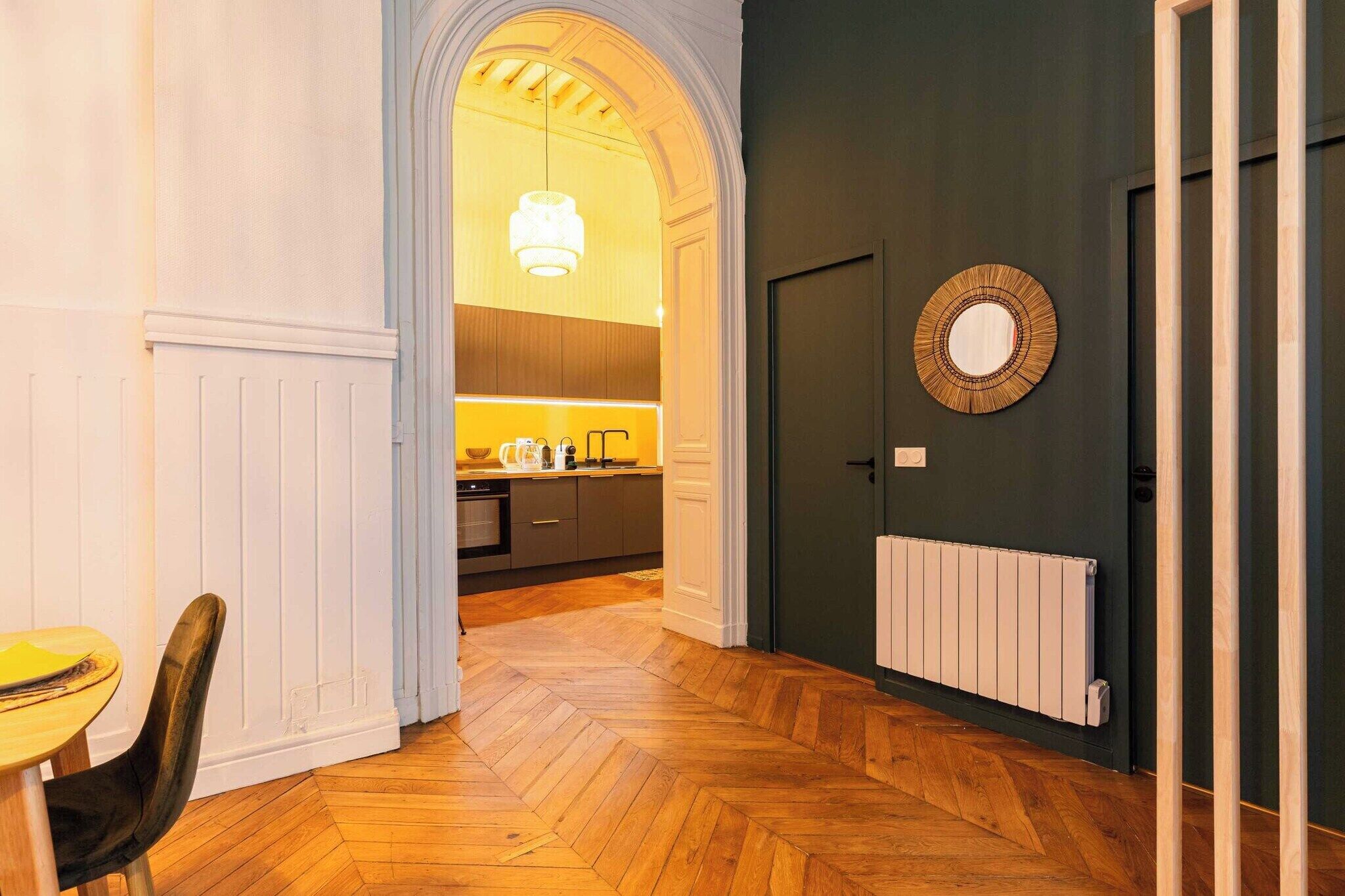 Un couloir doté d’un parquet en bois disposé en motif chevrons, un coin repas avec une table et des chaises, une cuisine équipée d’appareils modernes, et un mur sombre agrémenté d’un miroir rond.