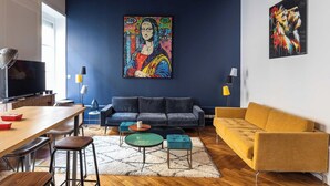 A modern living room with a dark blue accent wall, a large abstract painting, a sofa, a yellow armchair, and a dining area with a TV and bar stools. - DIFY Roi Lyon - Hotel de Ville (Lyon)