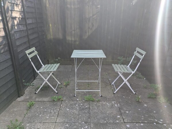 Un pequeño espacio al aire libre con una mesa de metal y dos sillas, frente a una valla de madera.