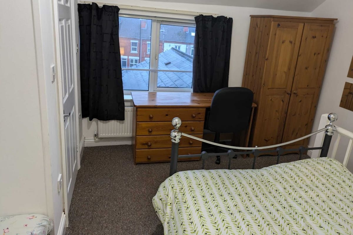A bedroom with a bed, a wooden desk, a chair, a wardrobe, and a window with curtains.