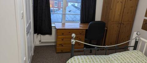A bedroom with a bed, a wooden desk, a chair, a wardrobe, and a window with curtains.