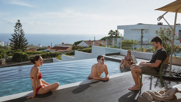 A group of people relaxing by a swimming pool with a view of the sea and buildings in the background.