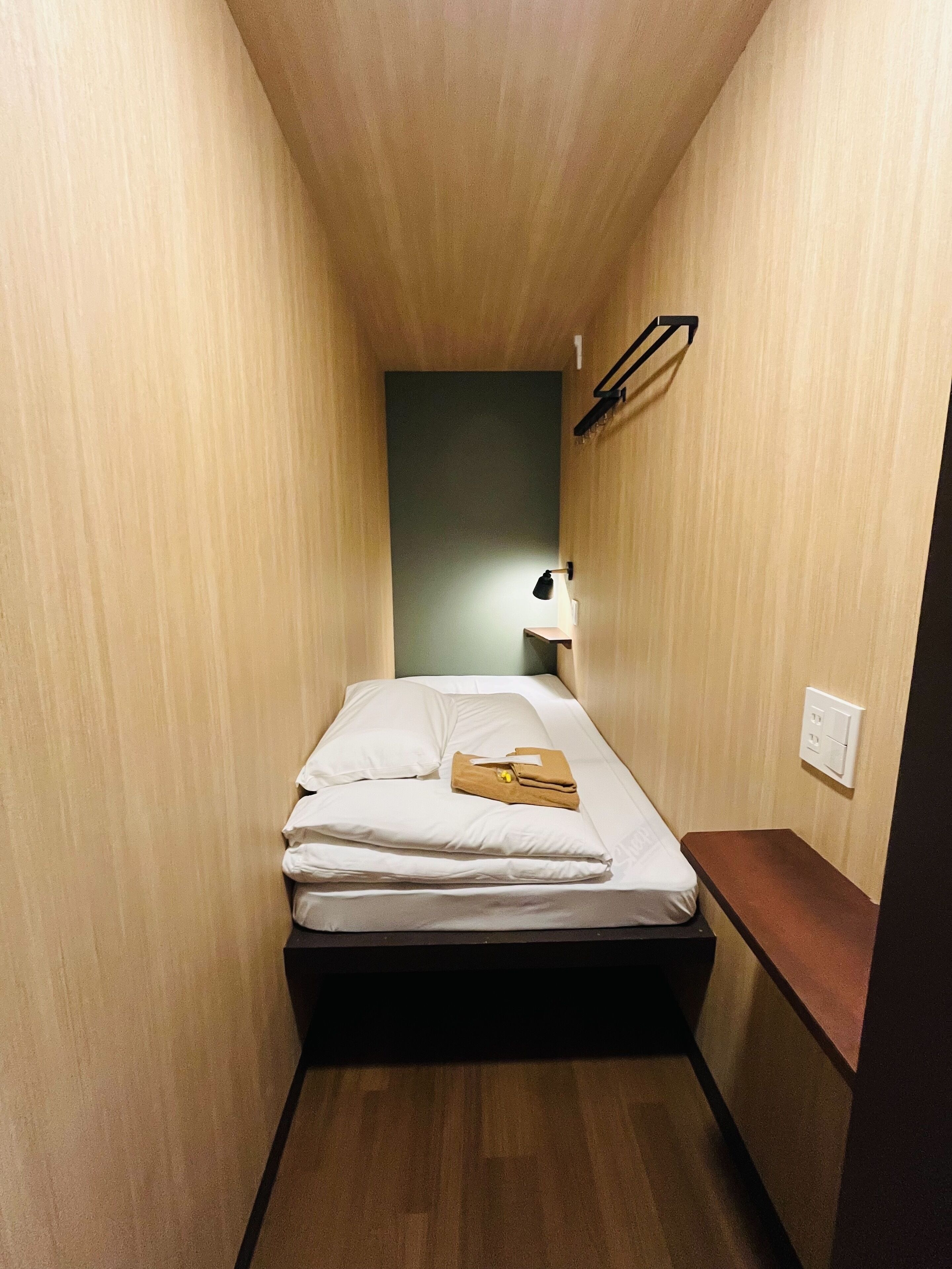 A narrow hotel room with a single bed, a bedside table, a lamp, and a wall-mounted shelf.