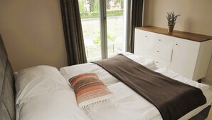 Ein ordentlich bezogenes Bett mit weißen Bettwäsche und einer braunen Tagesdecke, ein Nachttisch aus Holz mit einer Pflanze und ein Fenster mit Vorhängen.