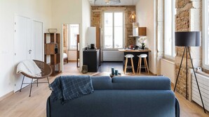 Ein modernes Wohnzimmer mit einem blauen Sofa, einem Korbstuhl, einer Küchenzeile mit Esstisch und Hockern sowie einem großen Fenster mit Aussicht.