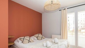 A bedroom with a bed, pillows, a bedside table, a lamp, and a large window with curtains.