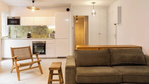 A modern kitchen with white cabinets, a microwave, and a coffee maker. There is a wooden chair and a small stool in the living area, and a brown sofa.