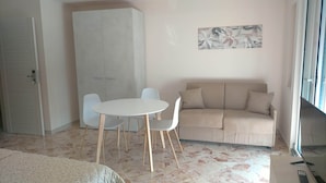 Un salon avec un canapé beige, une table ronde blanche et deux chaises, une armoire blanche et un tableau accroché au mur.