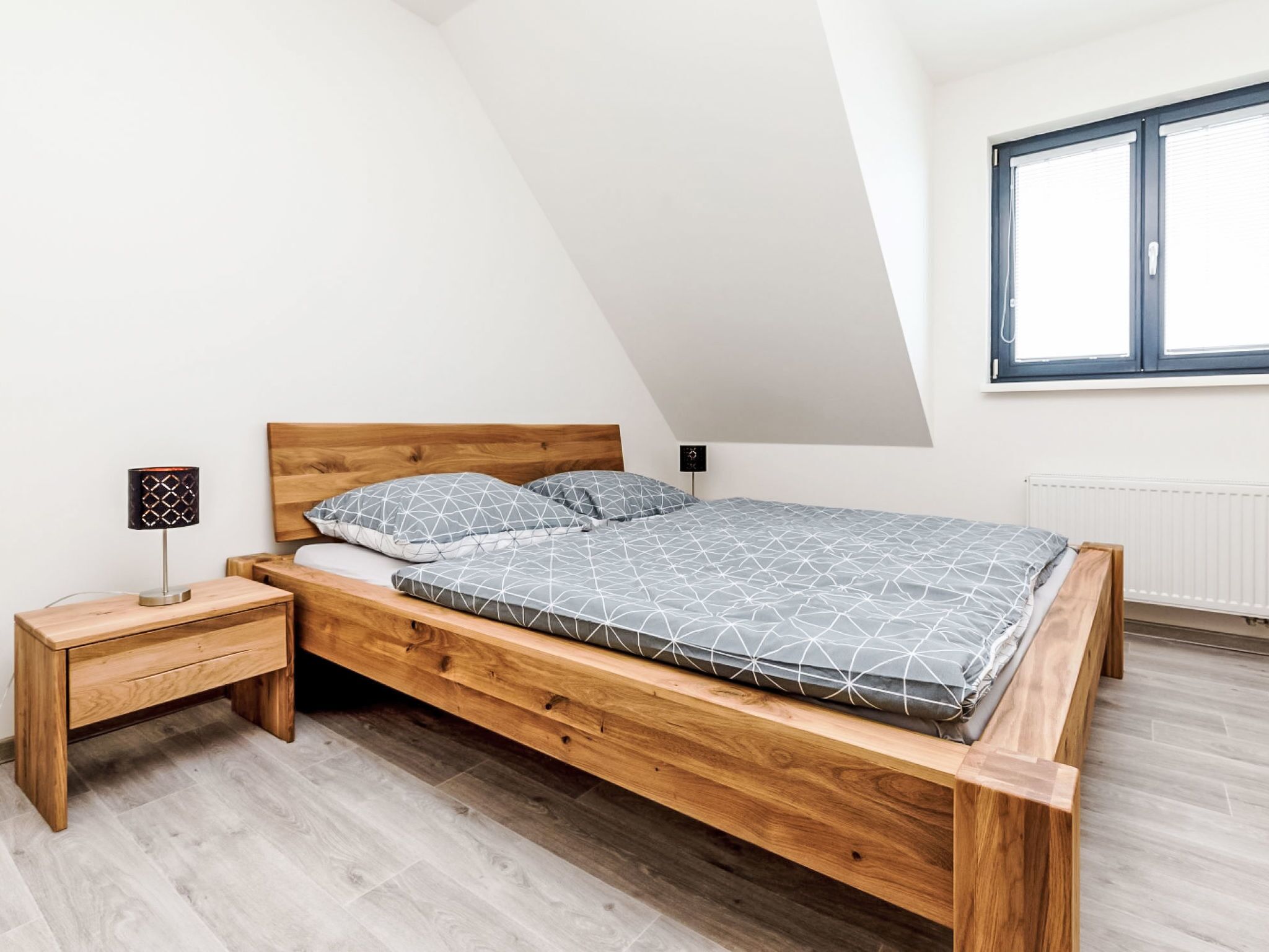 A bedroom with a sloped ceiling, a wooden bed frame, a bedside table, and a lamp.
