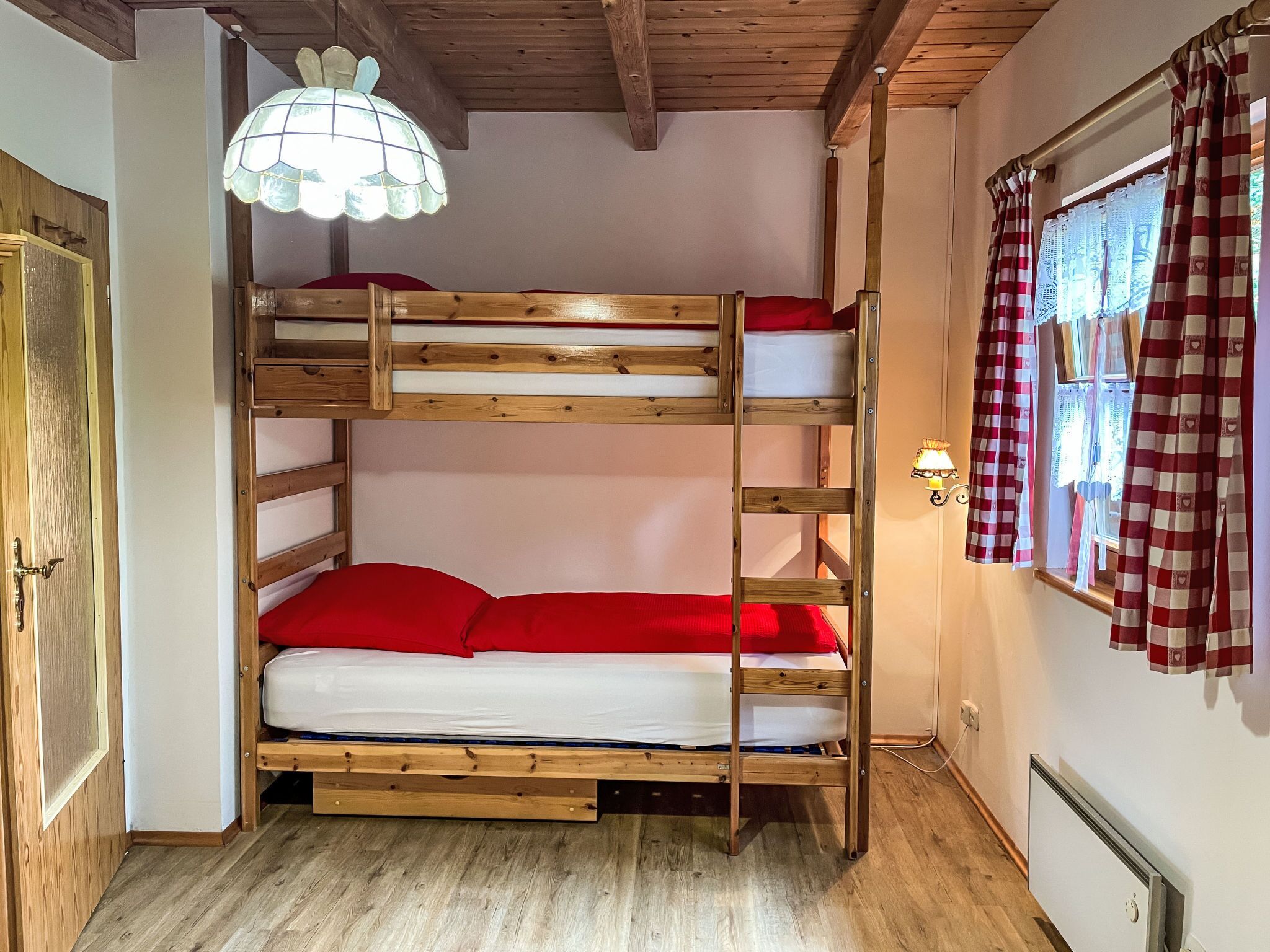 Un lit superposé avec une structure en bois et une literie rouge, une porte en bois, un luminaire fixé au mur et une fenêtre avec des rideaux à carreaux.