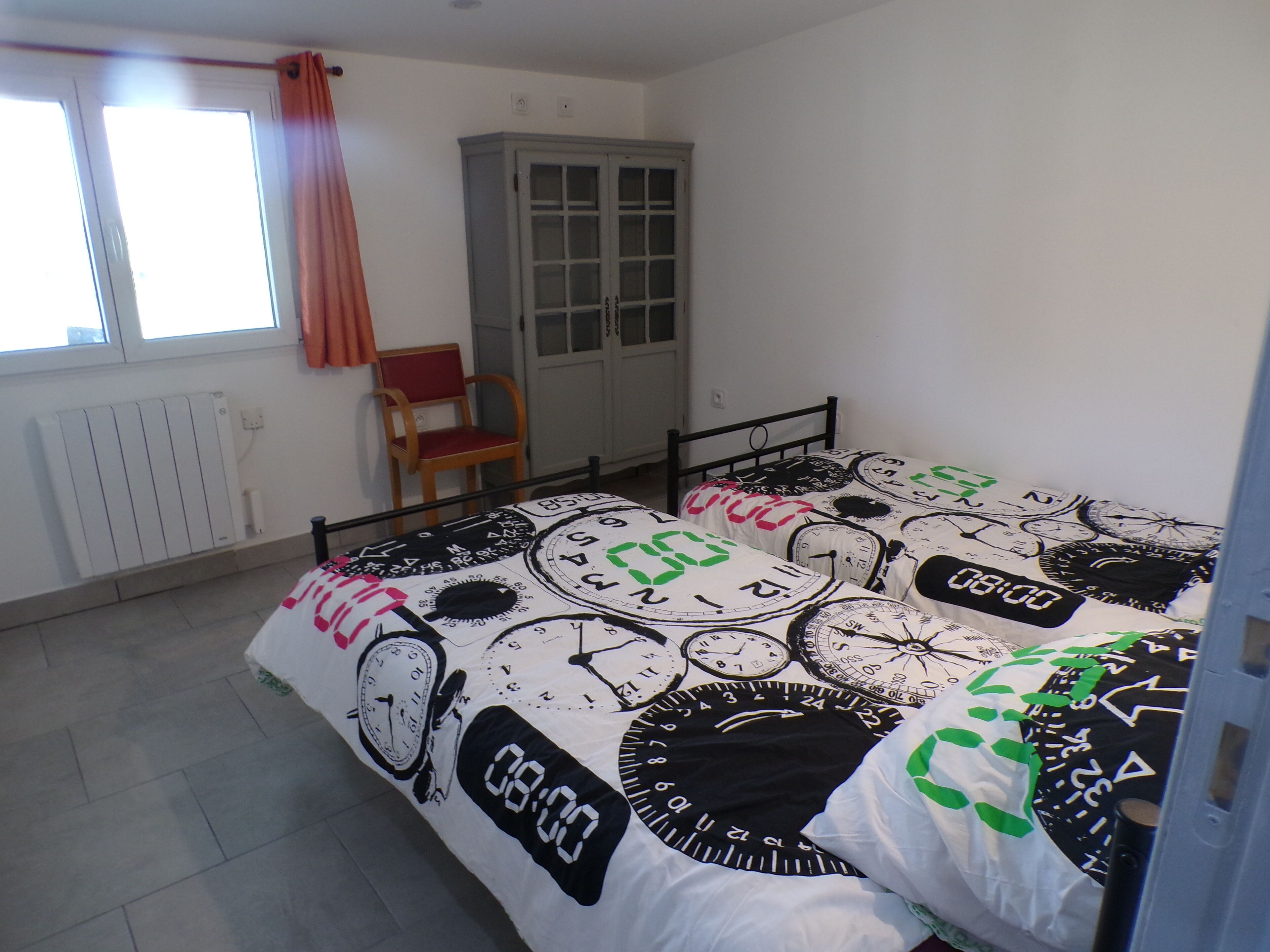 A bedroom with a bed featuring a clock-themed bedspread, a wooden chair, a wardrobe, and a radiator.