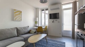 A modern living room with a grey sofa, a small table, and a TV mounted on the wall. There is a kitchen area with a fridge and a microwave. - DIFY Wilson - Villeurbanne (Villeurbanne)