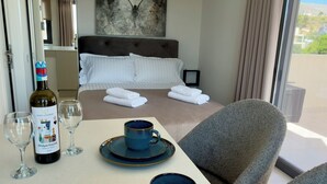 Une chambre d’hôtel avec un lit, une table à manger avec une bouteille de vin, des verres et une tasse, une chaise et une grande fenêtre.