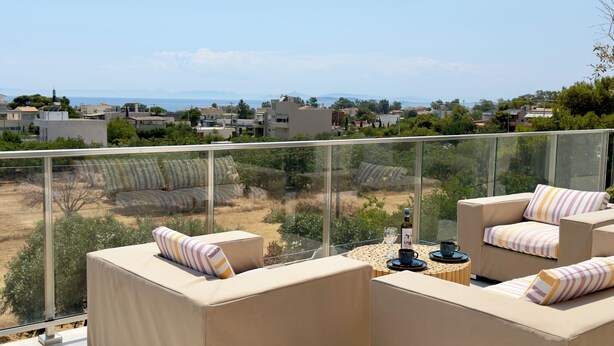 Un balcon doté d’une balustrade en verre, des canapés beiges avec des coussins à rayures, une petite table ronde avec une bouteille et des verres, et une vue sur un quartier résidentiel avec des maisons et des arbres.
