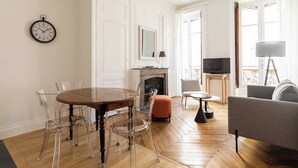 Een woonkamer met houten vloer, een ronde eettafel met transparante stoelen, een open haard, een televisie en een bank.